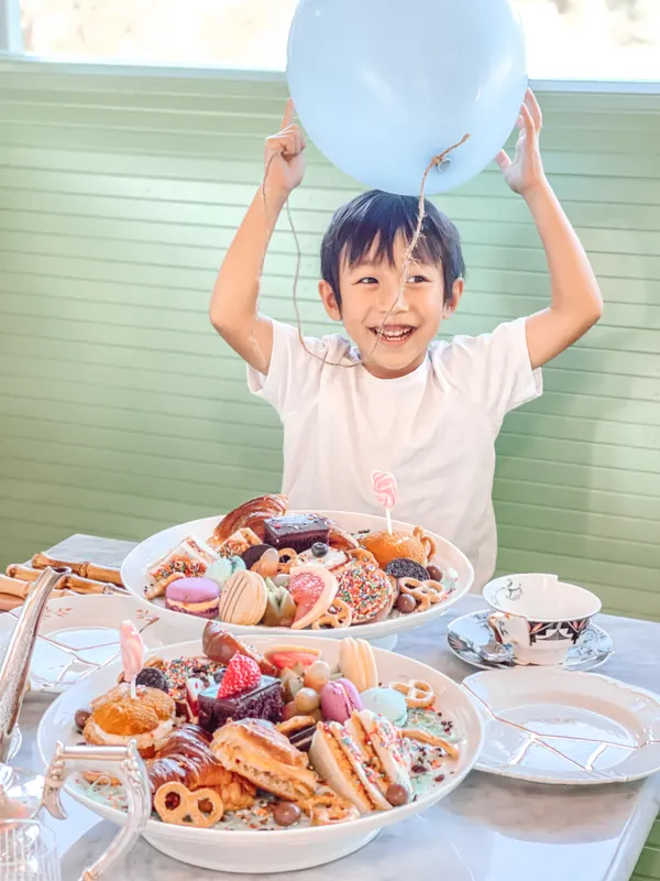 A boy holding a balloon overhead behind a table of high-tea treats
