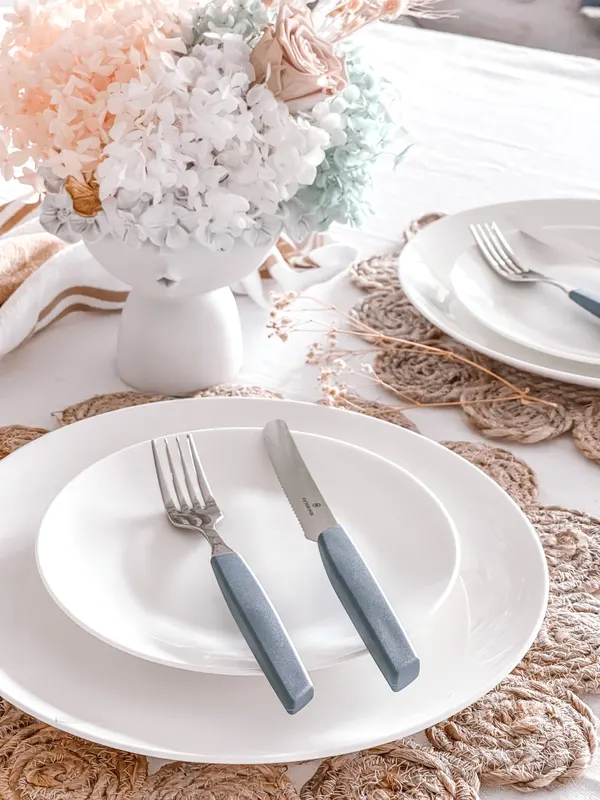 A dinner setting with a white plate, blue-handled cutlery and a woven placemat